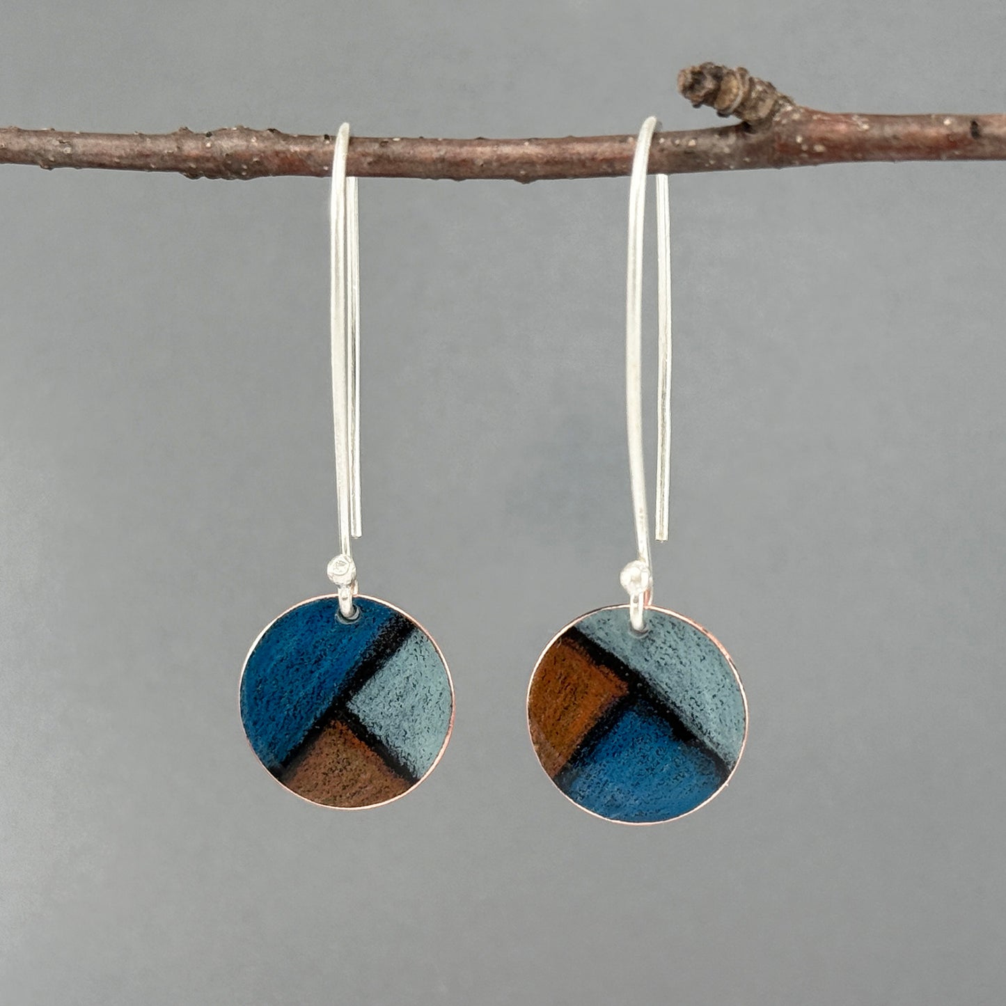 Pair of round earrings with blue and dark orange design hanging on a branch against a gray background