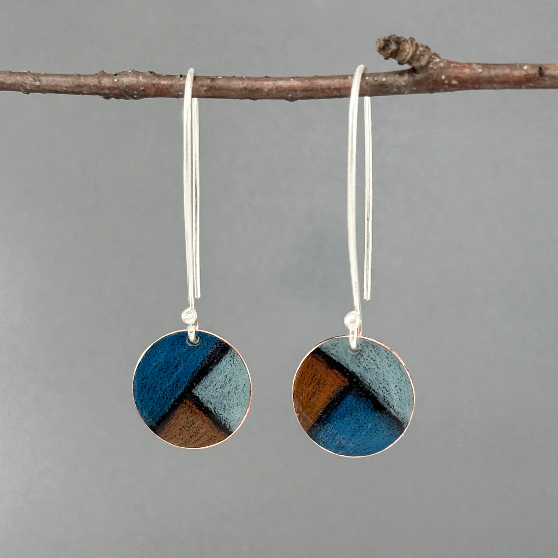 Pair of round earrings with blue and dark orange design hanging on a branch against a gray background